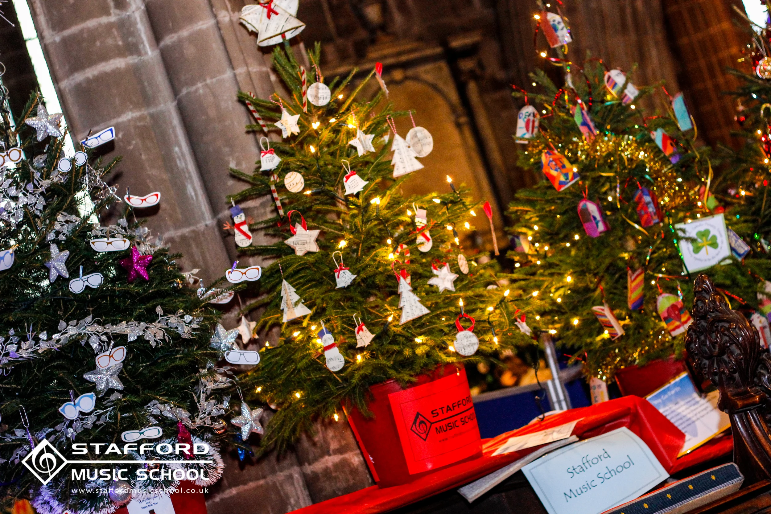 Three decorated Christmas trees with ornaments and lights at Stafford Music School, displayed against a brick wall, with some packages and signs visible at the bottom.