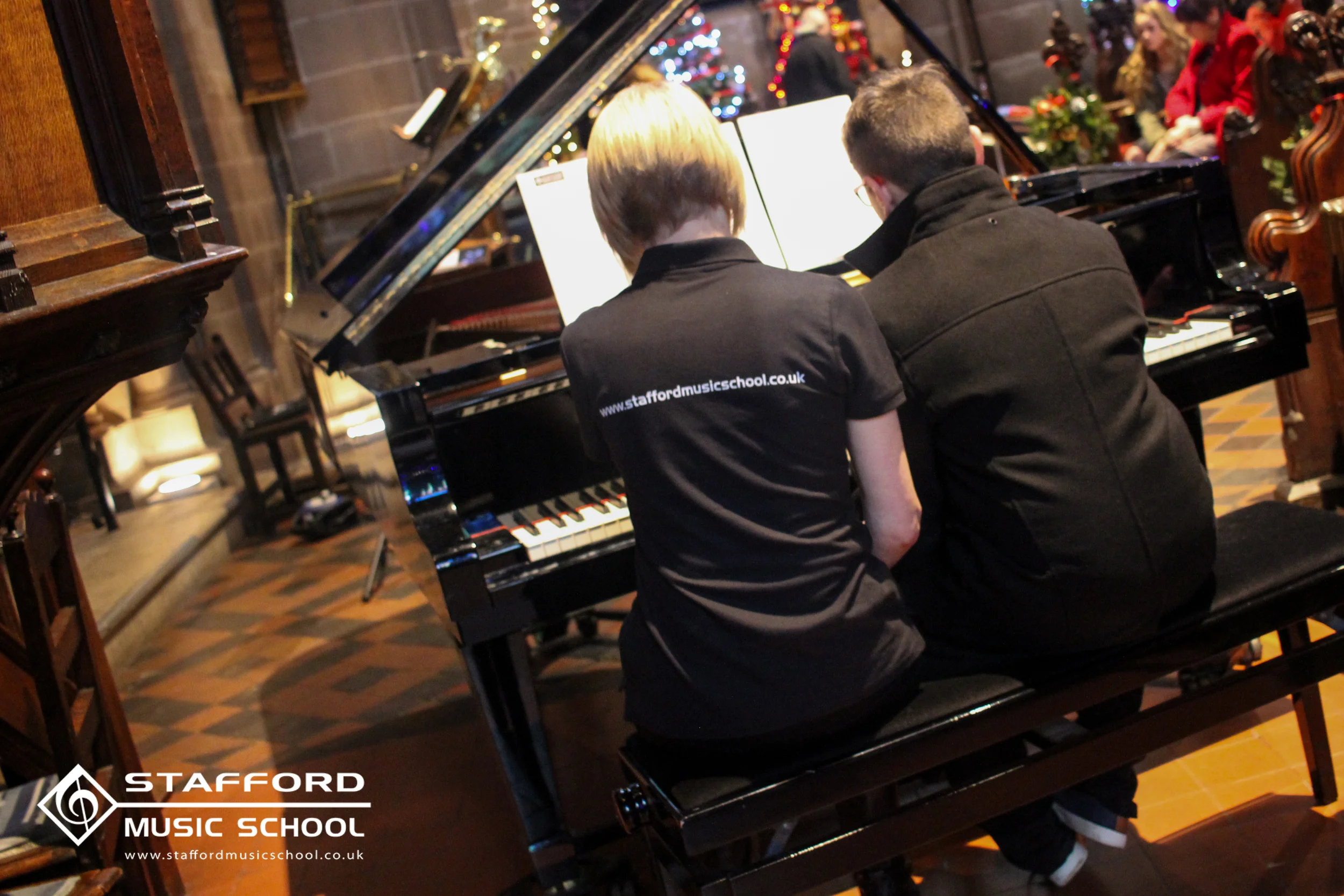 Two students are sitting at a grand piano, playing music together in a decorated, festive room with Christmas trees and ornaments. The students are facing away from the camera, and the scene includes other people in the background, likely at a holida