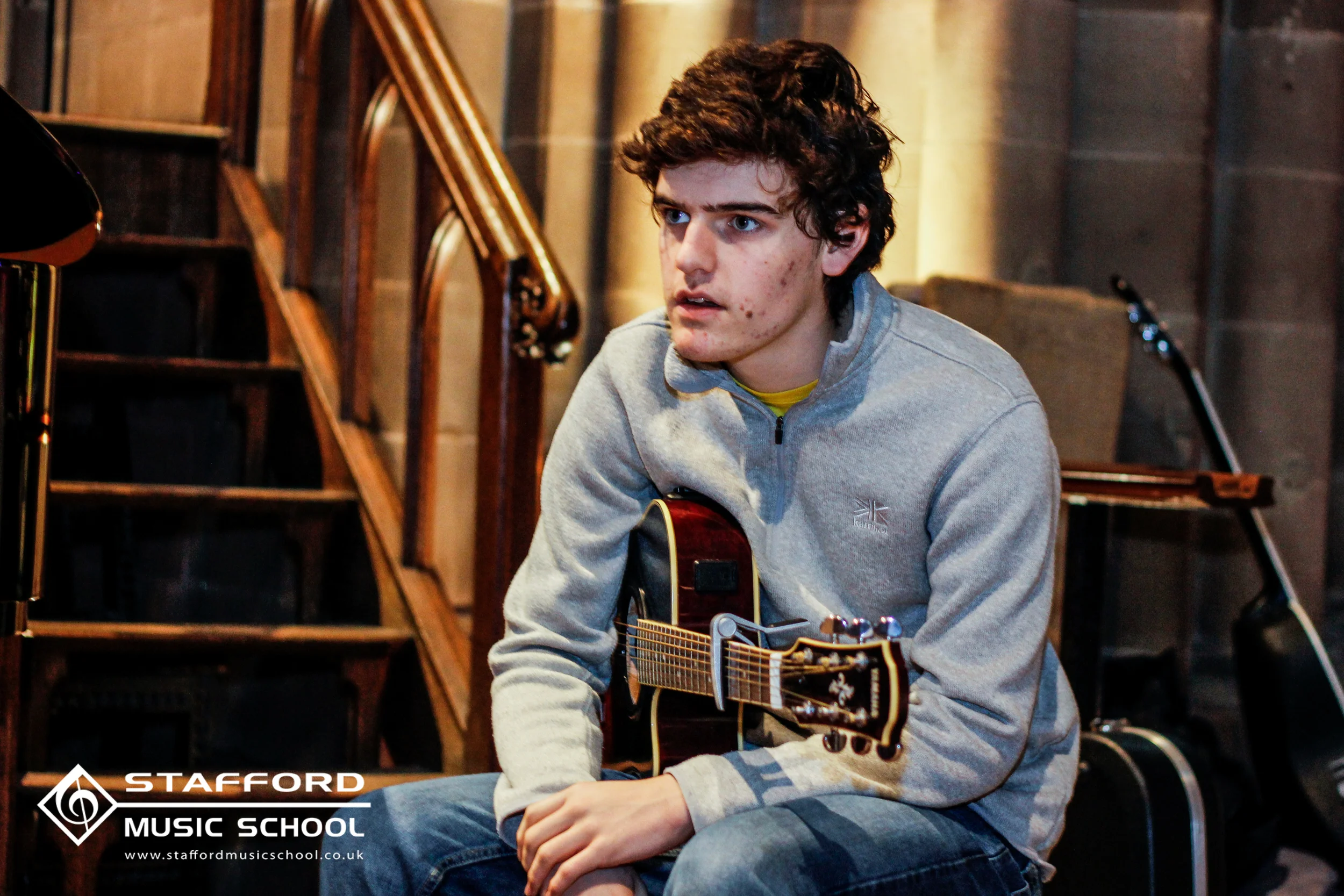 Young man with dark curly hair and acne playing an acoustic guitar in a music studio, wearing a gray zip-up sweatshirt and blue jeans, with stairs and recording equipment in the background.