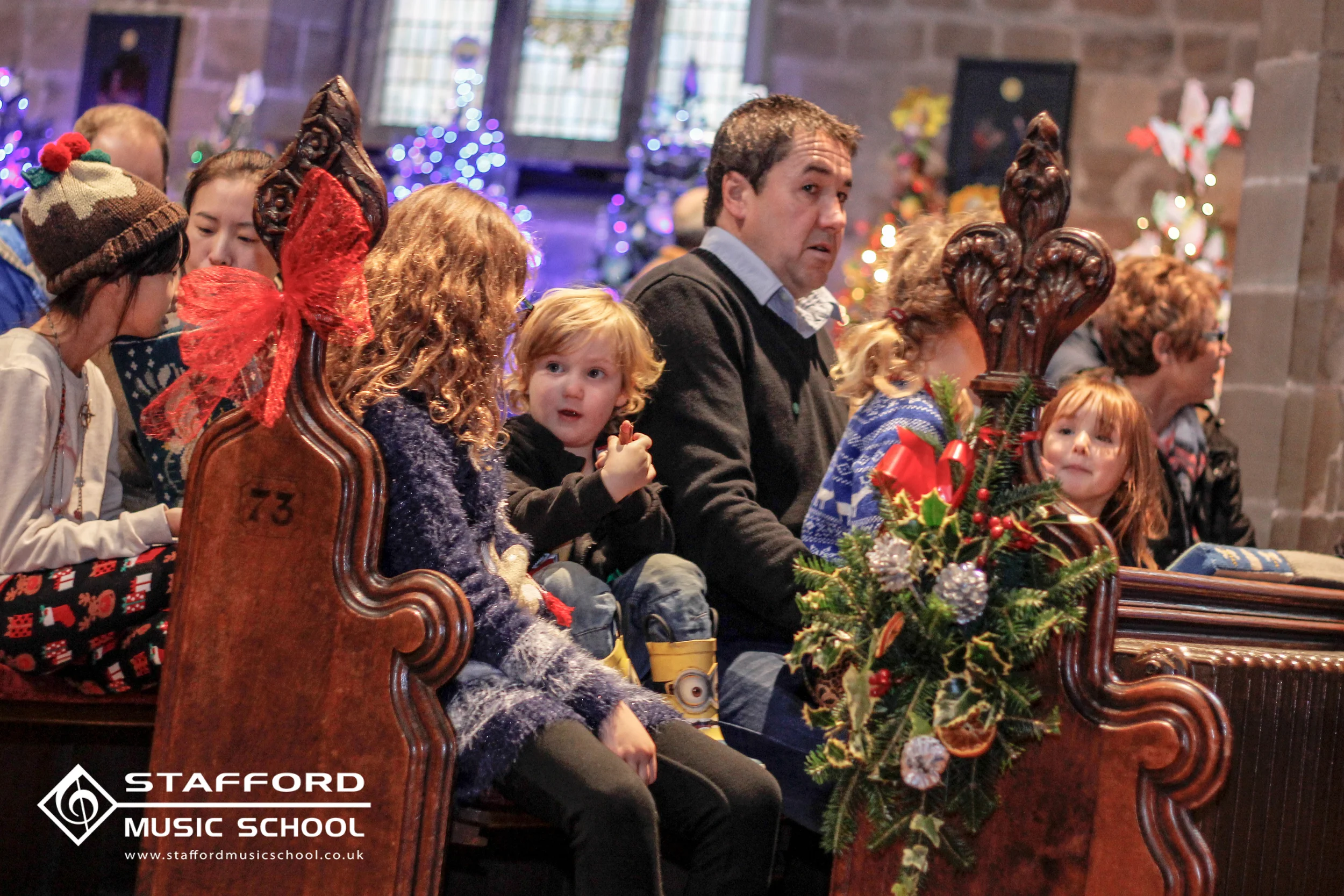 Children and adults sitting on wooden pews in a decorated church for a Christmas event, with holiday decorations, Christmas trees, and holiday lights in the background.