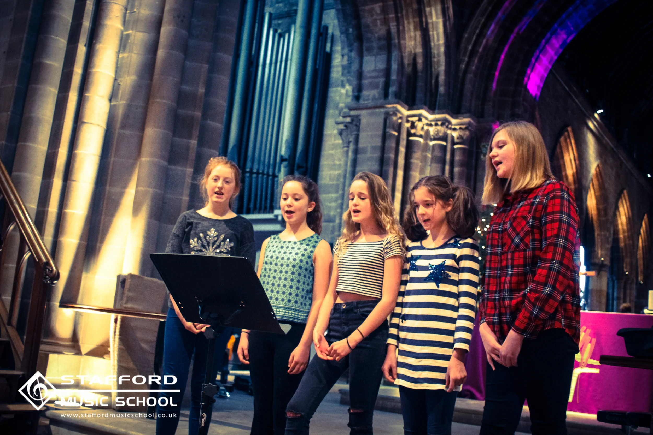 Group of children singing together in a music school, standing in a semi-circle and facing a music stand, in a church or large hall with stained glass windows and stone columns.