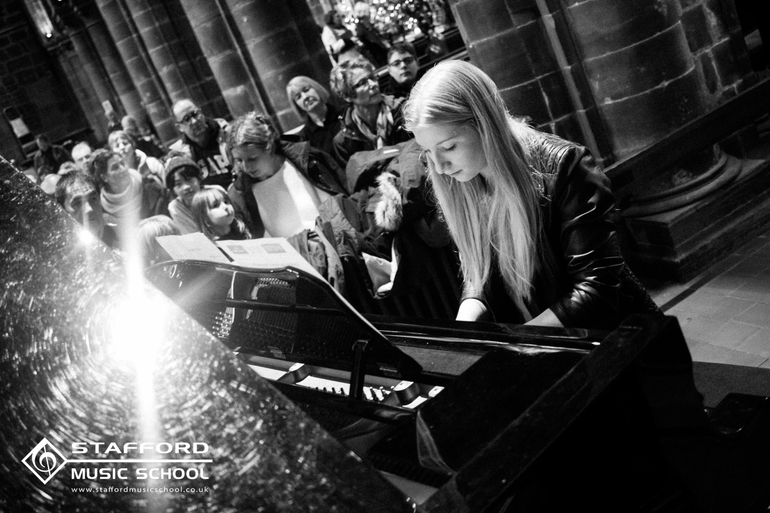 A group of people, including children and adults, sitting inside a church or similar venue, watching a young woman playing the piano. There is a logo in the bottom left corner for Stafford Music School.