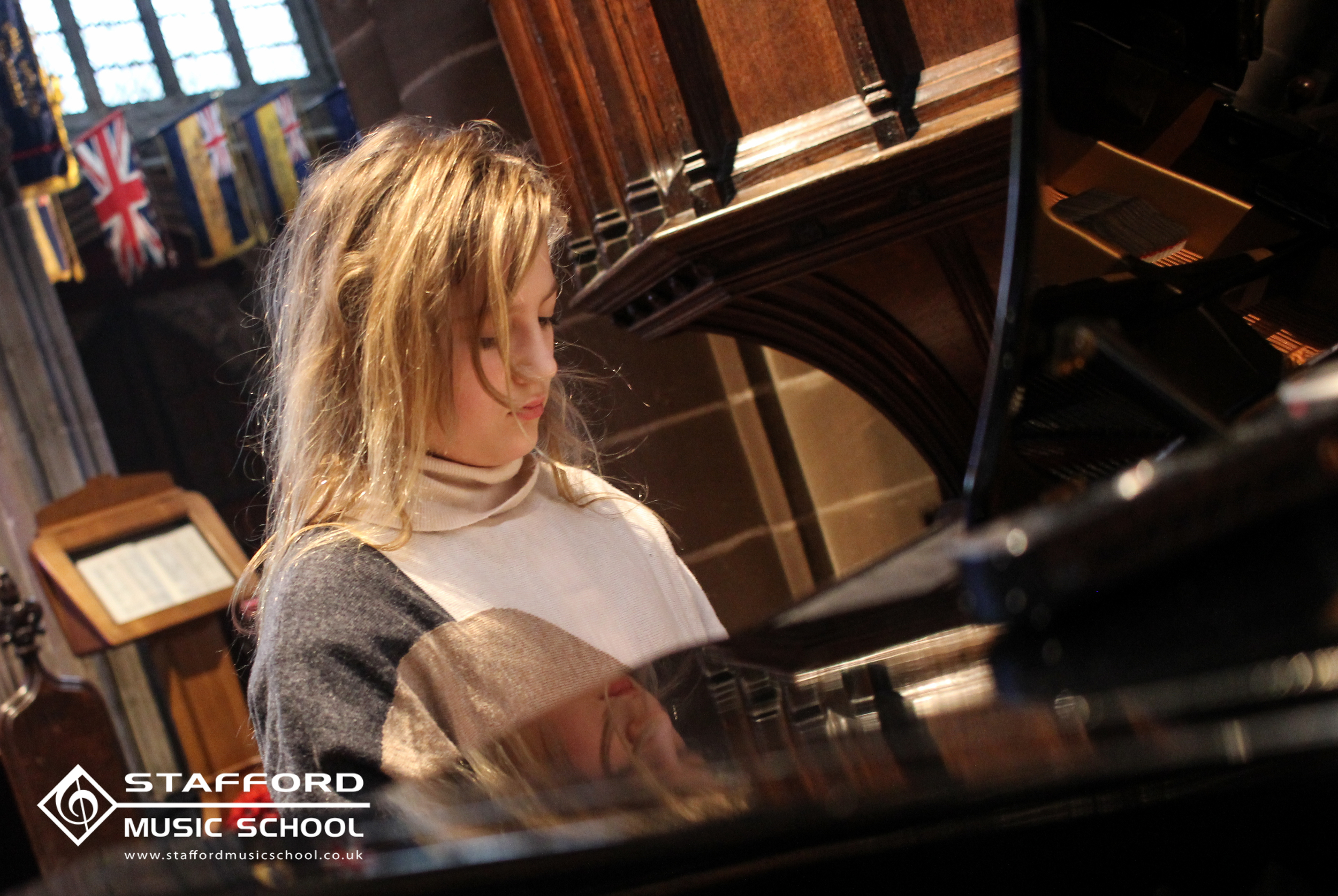 A young woman with blonde hair playing a grand piano in a music school. Flags are hanging from the ceiling in the background.