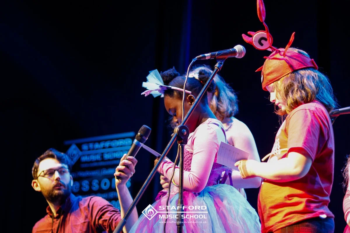 Children dressed in colorful costumes performing or practicing on stage with microphones at Stafford Music School.