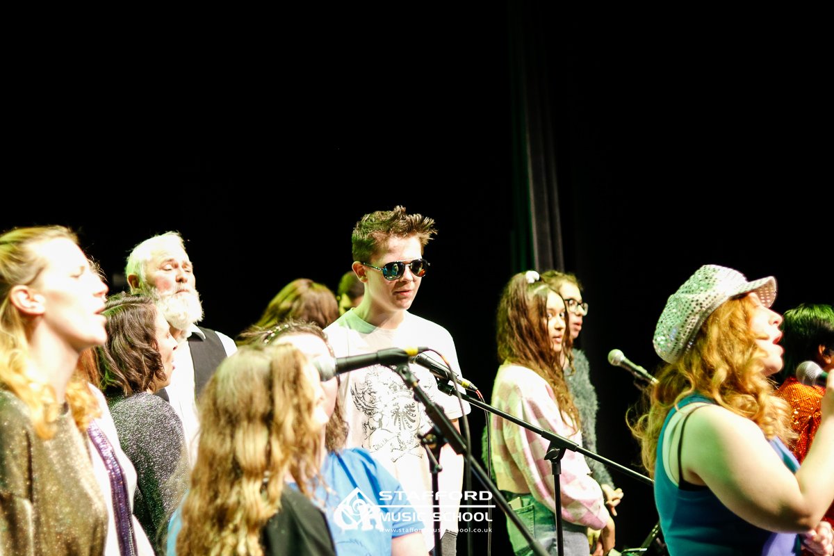 Group of diverse young people singing or performing on stage, with microphones, against a dark background at Stafford Music School.