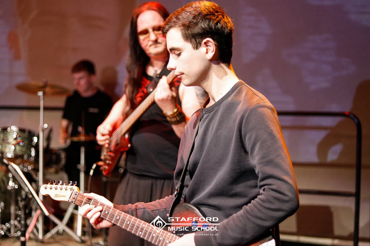 Young man in a gray sweatshirt playing an electric guitar during a band performance with a drummer and a bassist in the background at Stafford Music School.