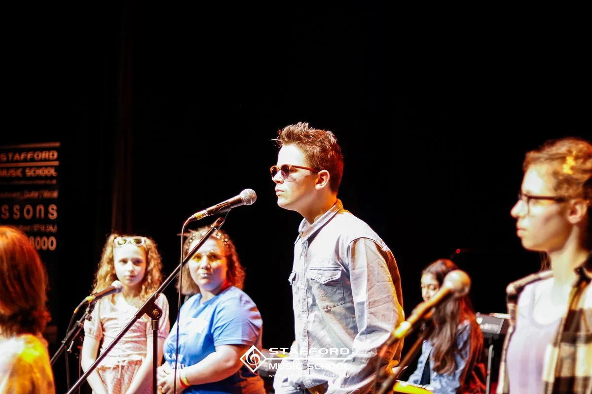 Group of young students on stage at Stafford Music School, with one boy wearing sunglasses and speaking into a microphone, while others look on, in a dark room with stage lighting.