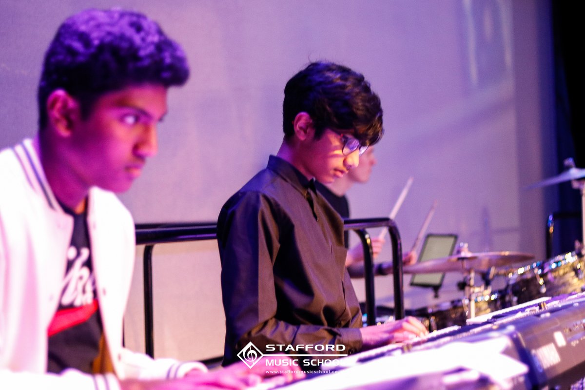 Three young musicians playing electronic keyboards on stage at Stafford Music School, with a purple background.