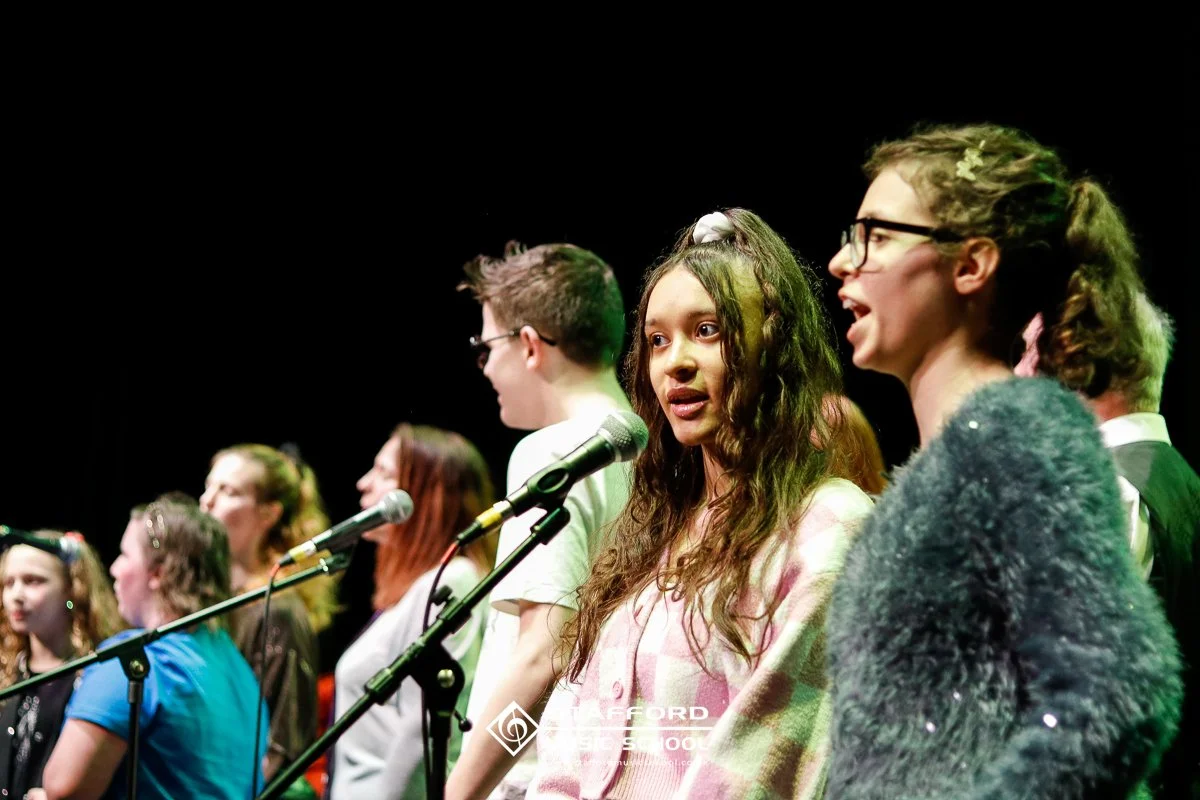 A group of young students, standing on stage in front of microphones during a school event, with some wearing glasses and casual attire, and a dark background.