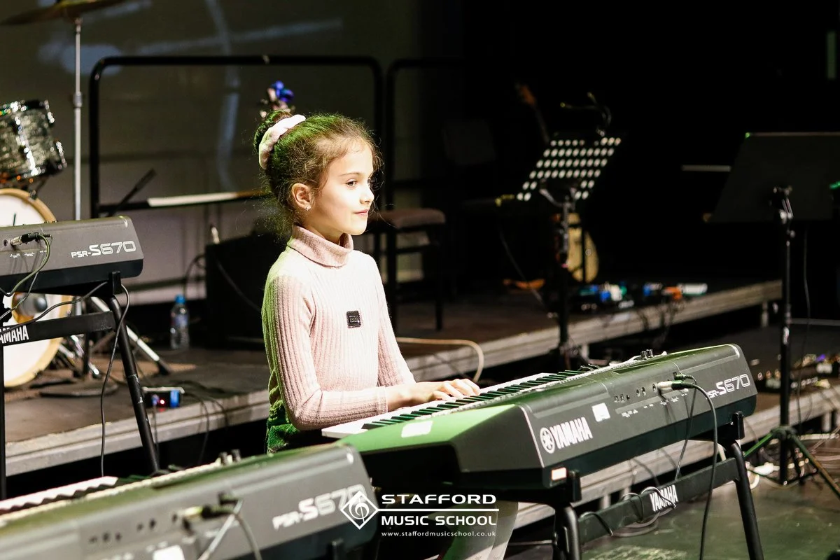 Young girl sitting at a Yamaha keyboard on stage, practicing or performing, with music stands and drums visible in the background at Stafford Music School.