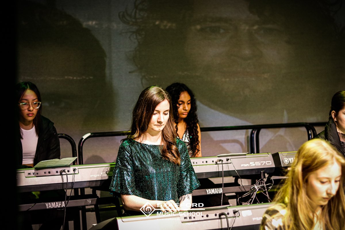 Group of young women playing Yamaha keyboards on stage, with one in the foreground and a large projected face on the screen behind them.