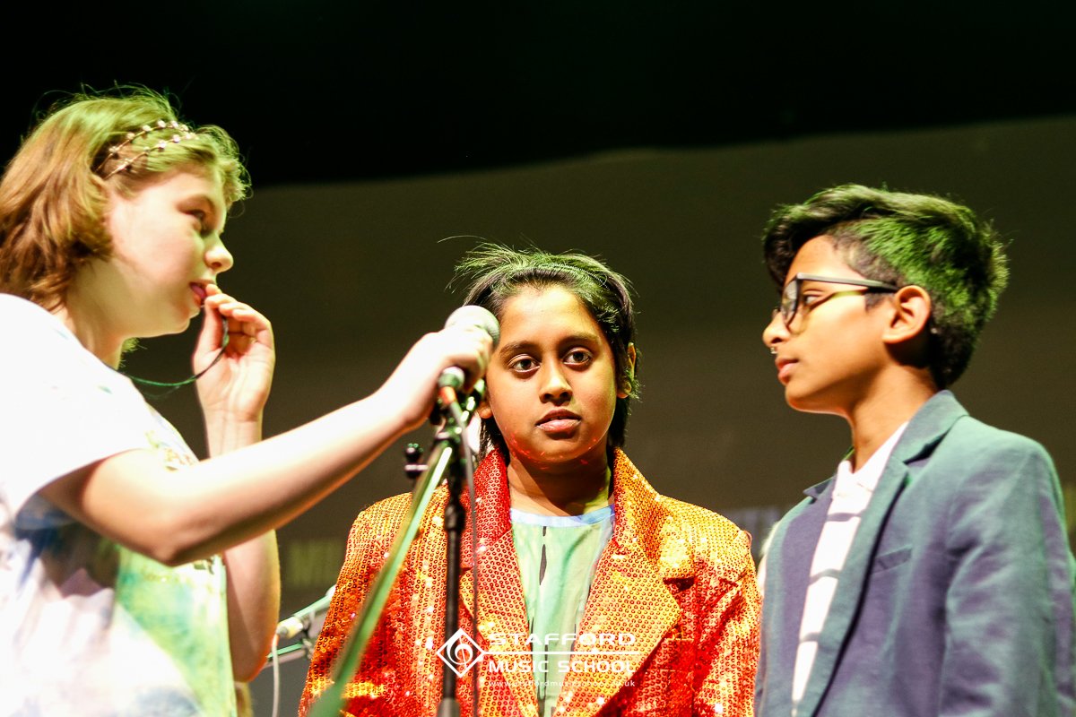 Three young people on stage, two girls and one boy, engaged in an activity possibly related to music or performance, with a microphone in front of the girl in the middle.