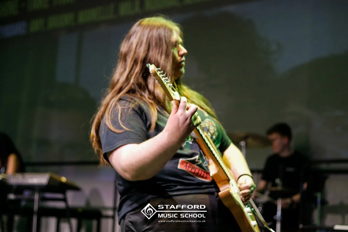 A person with long, reddish-brown hair playing an electric guitar at Stafford Music School.