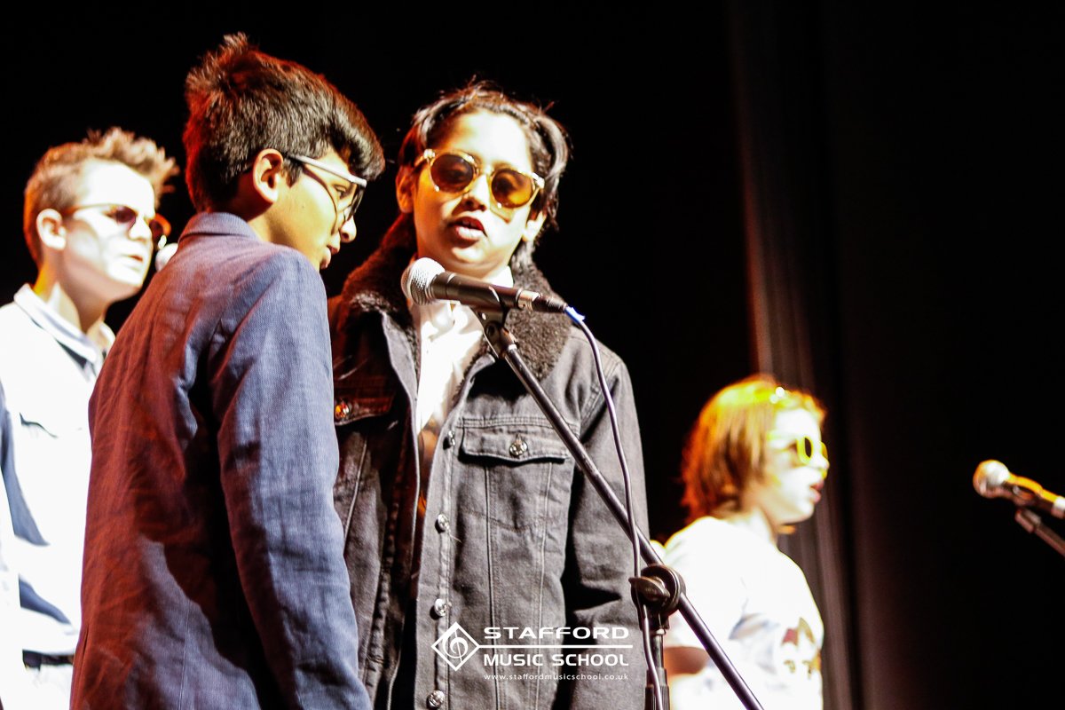 Group of children performing on stage at Stafford Music School, with one girl wearing sunglasses in front of a microphone.
