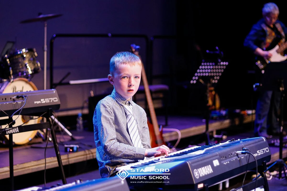 Young boy playing a Yamaha keyboard during a music performance, with other musicians playing instruments in the background at Stafford Music School.
