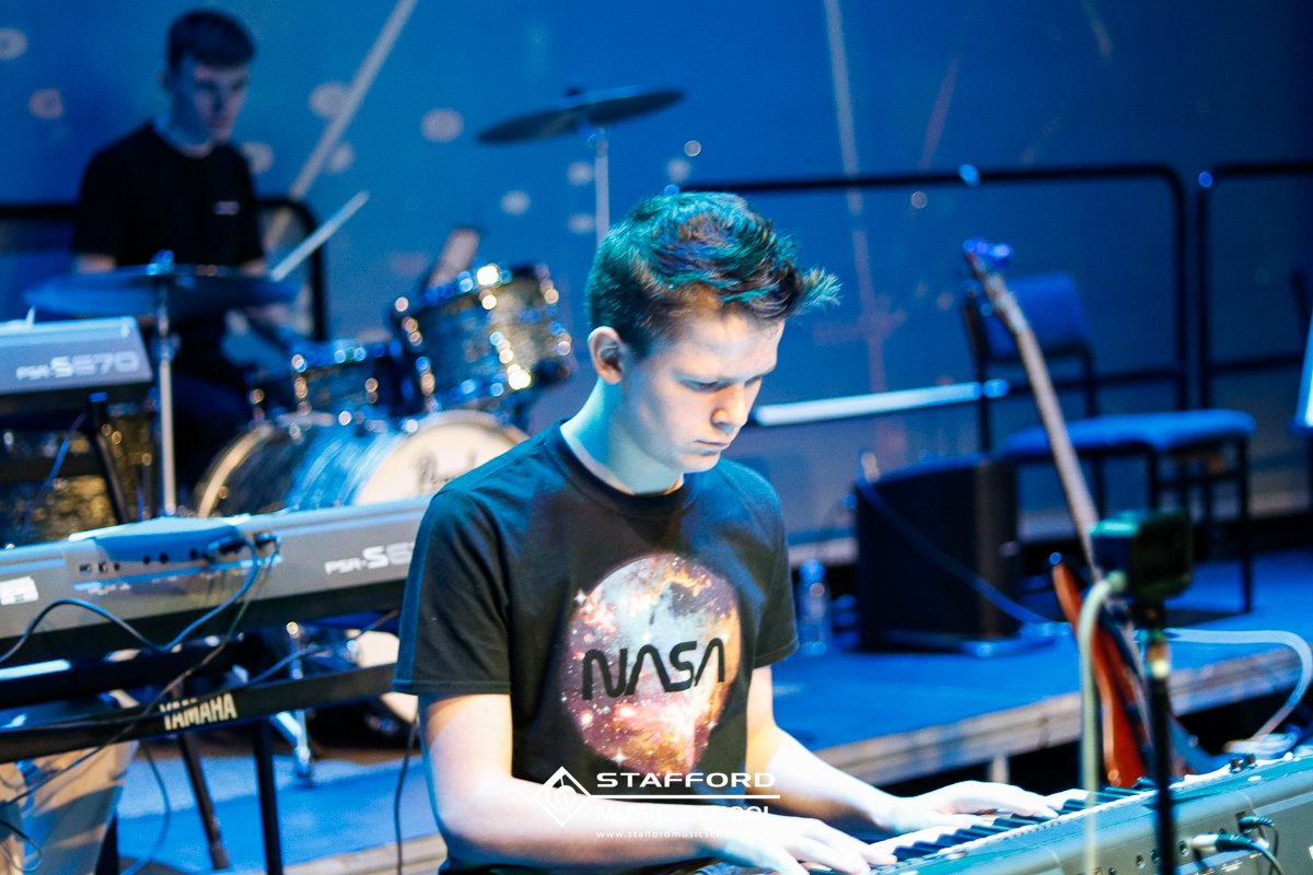Young man wearing a NASA T-shirt playing a keyboard on stage during a performance with a drummer in the background.