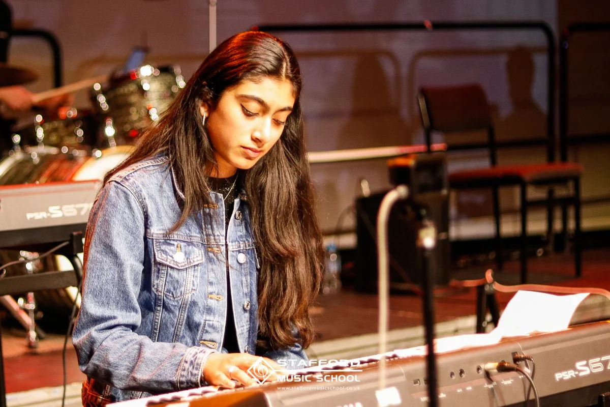 Young woman with long dark hair wearing a denim jacket playing a keyboard in a music studio or rehearsal space.