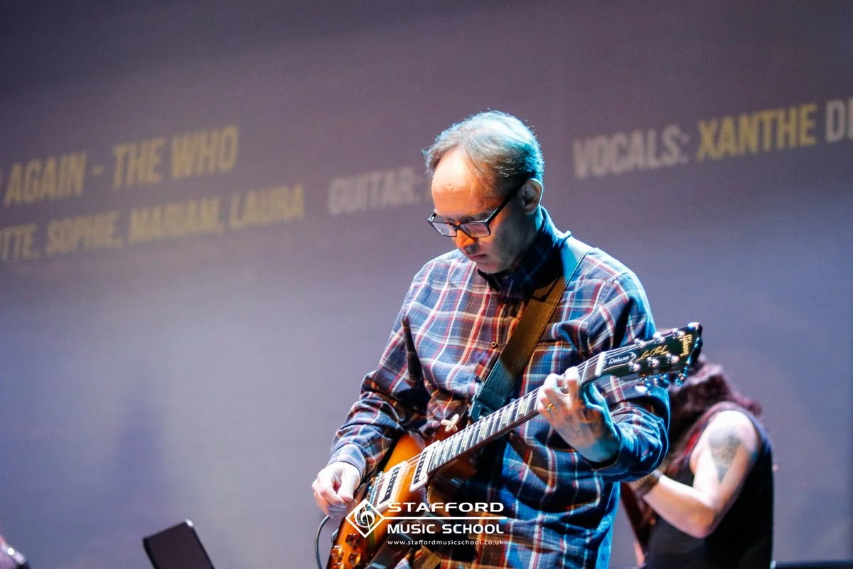 Man playing an electric guitar while wearing glasses and a plaid shirt during a music event.