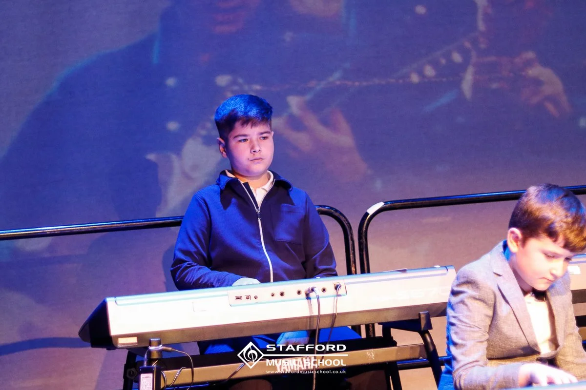 Two boys performing on stage, one playing a keyboard and the other sitting nearby, with a dark background.