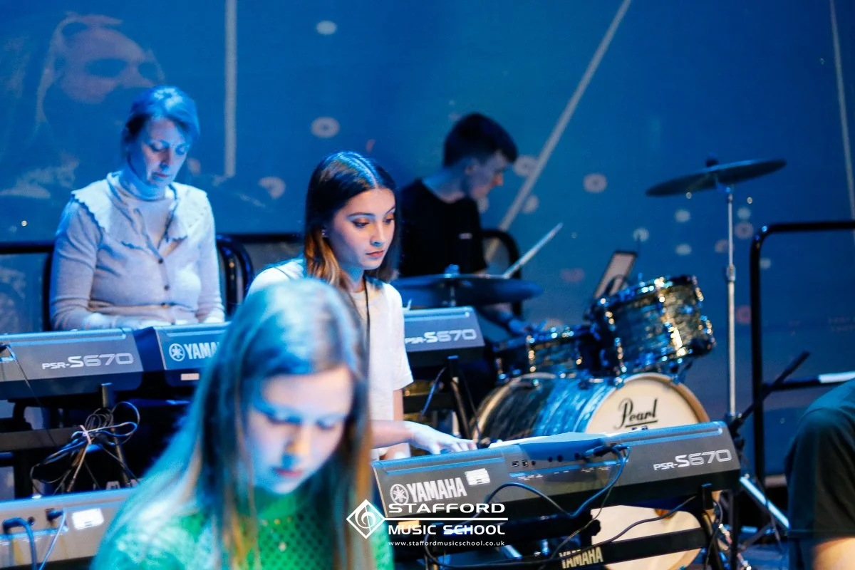 Young women practicing keyboard and drums during a band rehearsal or music class at Stafford Music School, with musical instruments and equipment visible.