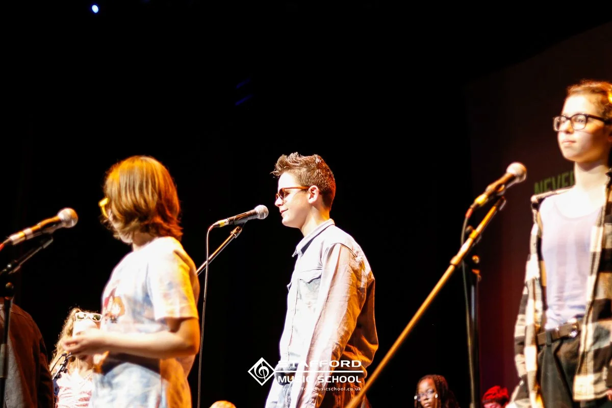 Three teenagers standing on stage in front of microphones at a music school performance, with dark background and stage lighting.
