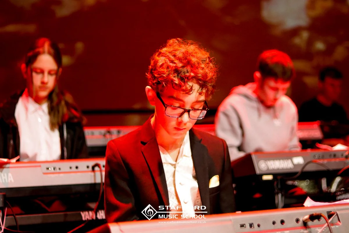 Young male with curly hair and glasses, playing a keyboard during a music performance at a music school.