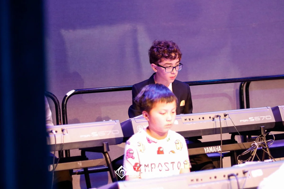 Two young boys sitting at a Yamaha keyboard, one in a suit and glasses and the other in a white shirt with colorful patterns, during a performance or rehearsal on stage.