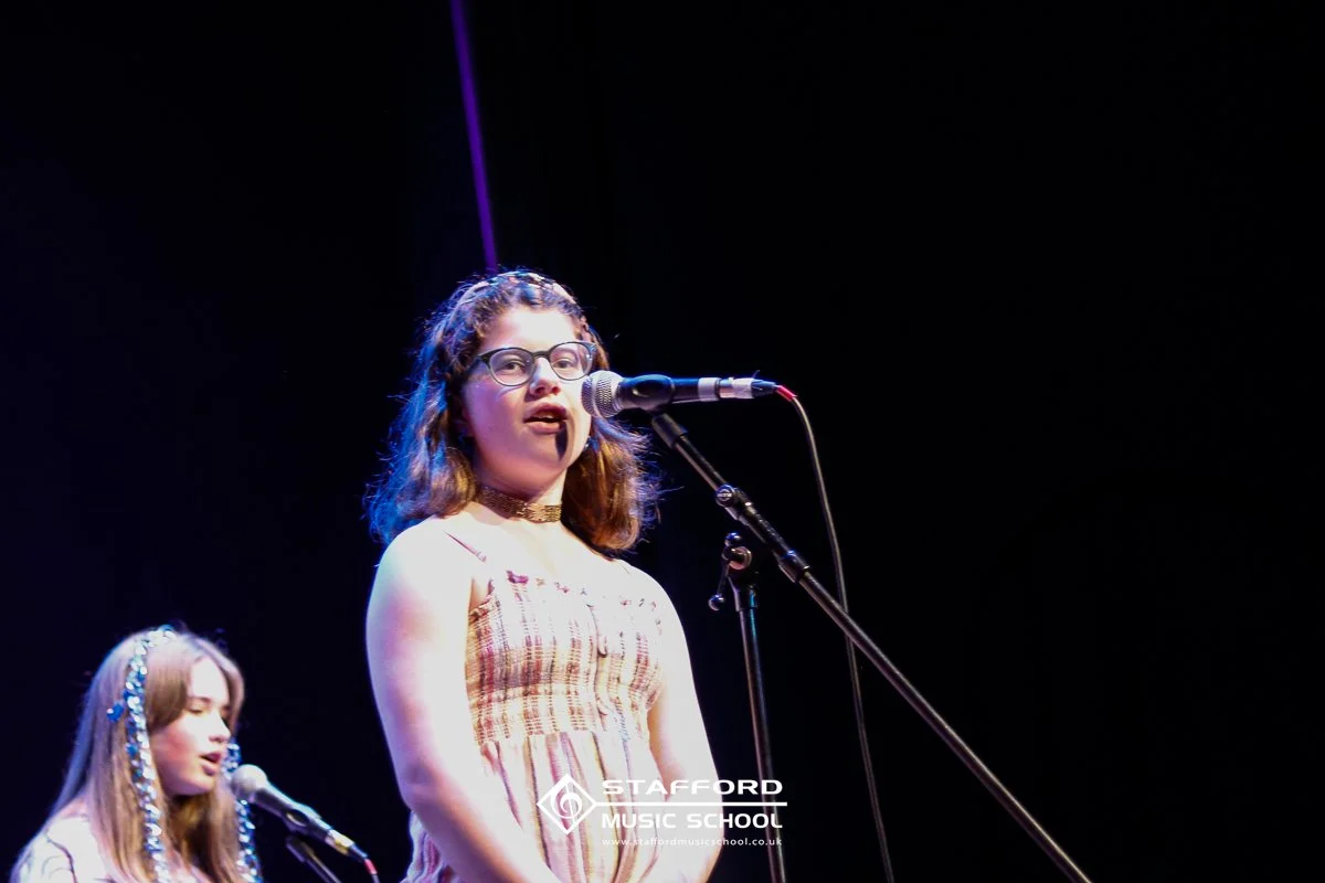 Two young girls singing on stage at Stafford Music School. The girl in front wears glasses, a striped dress, and a choker. The girl behind has long hair with decorative accessories and is also singing.