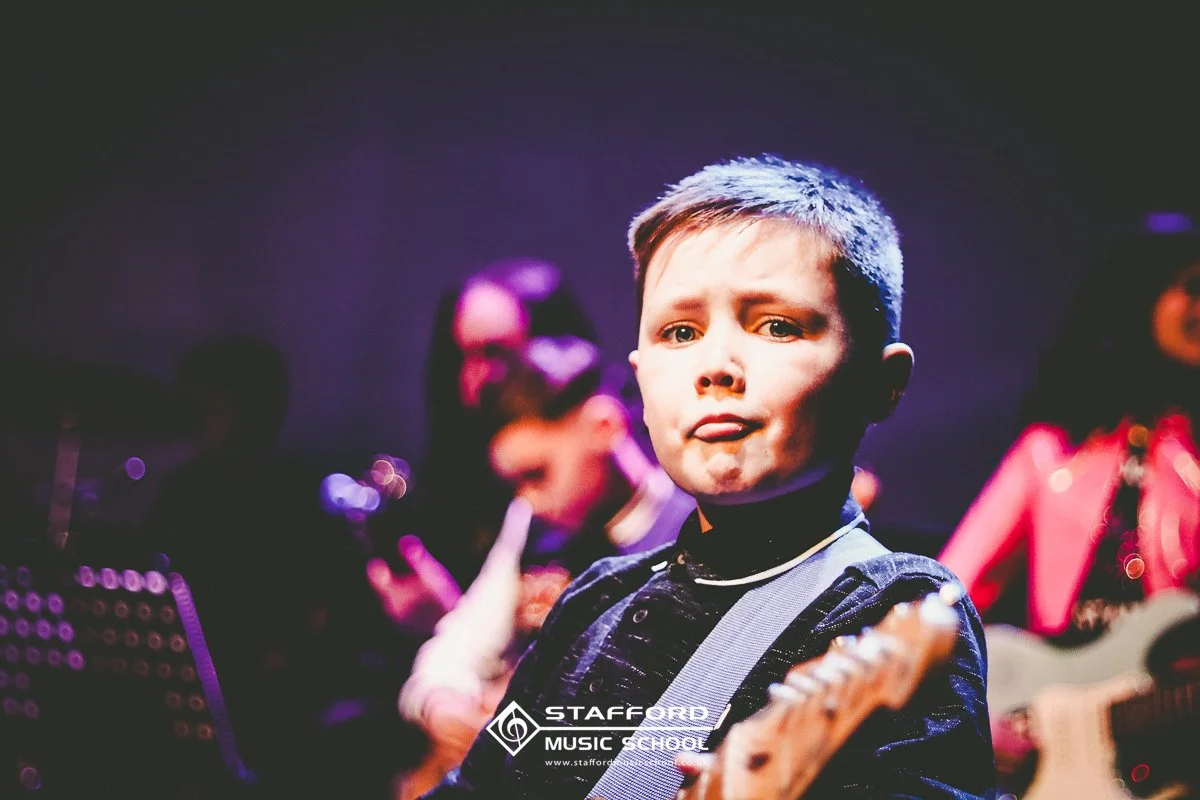 A young boy at a music school performance, standing with a guitar. He has a serious expression, and other children are in the background, some playing guitars. The setting appears to be a stage with dark lighting and colorful accents.