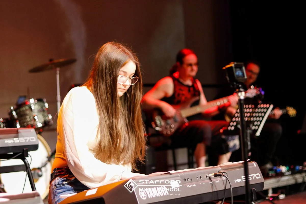 A young woman playing a keyboard on stage, with two other musicians in the background, one playing a bass guitar, and another with an electric guitar. The scene appears to be a live music performance.