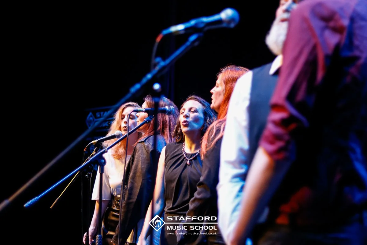 Group of women singing on stage with microphones, colorful stage lighting, and a black background, at Stafford Music School.