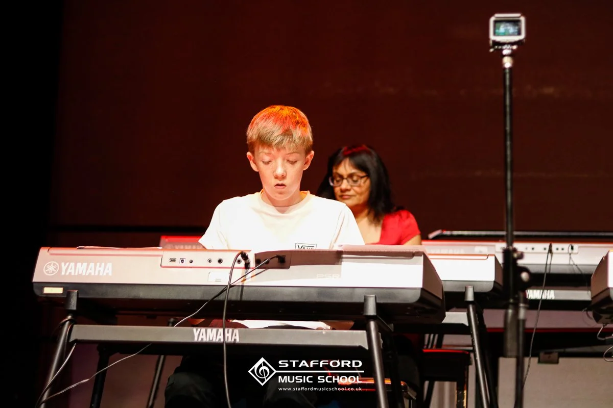 A young boy playing a Yamaha keyboard on stage, with a woman in the background and a microphone stand to the right, at Stafford Music School.