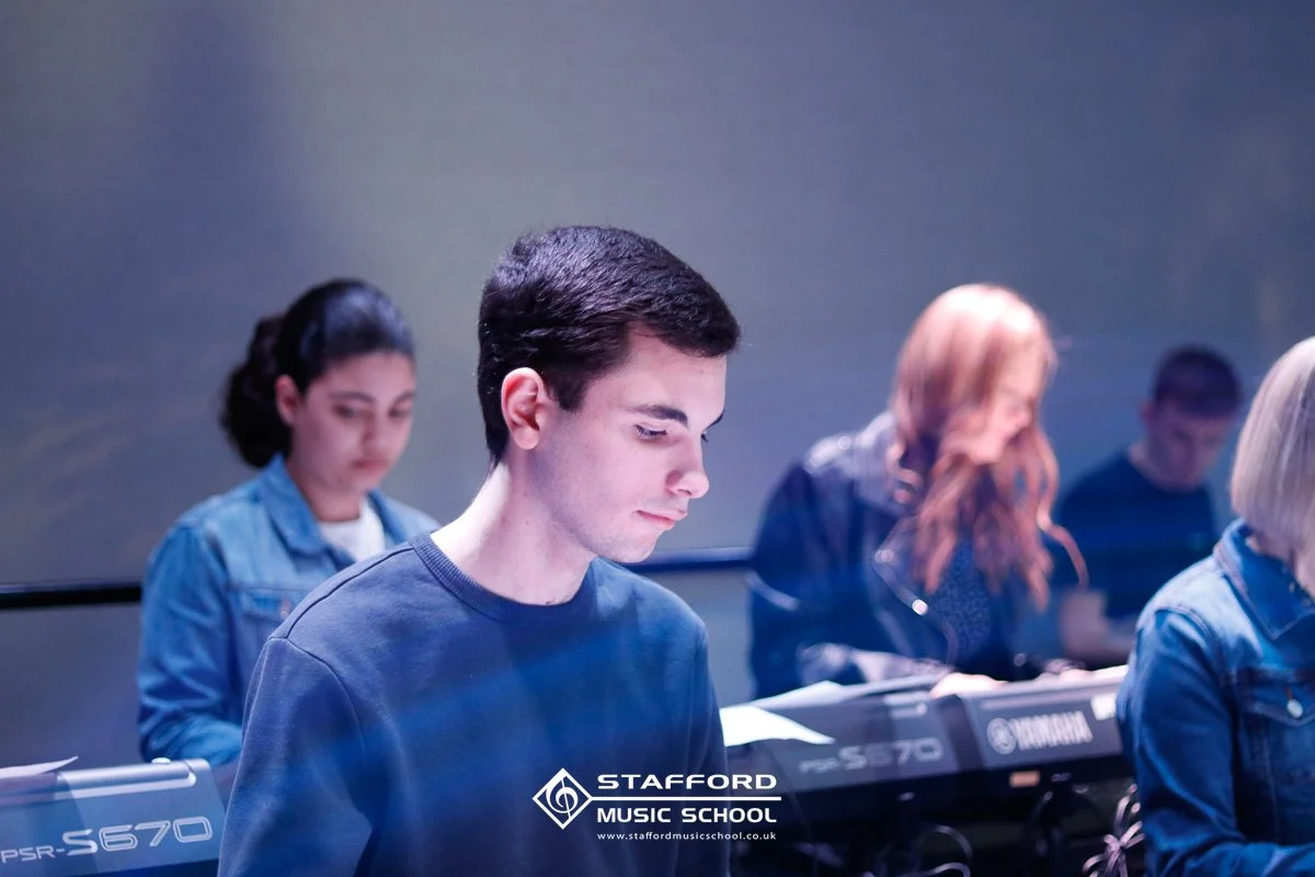 Young man in a music classroom, playing a keyboard with other students in the background, Stafford Music School logo visible.