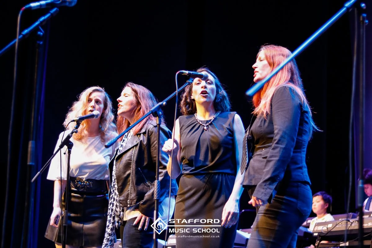 Four women singing on stage with microphones, accompanied by musicians, under stage lighting at Stafford Music School.