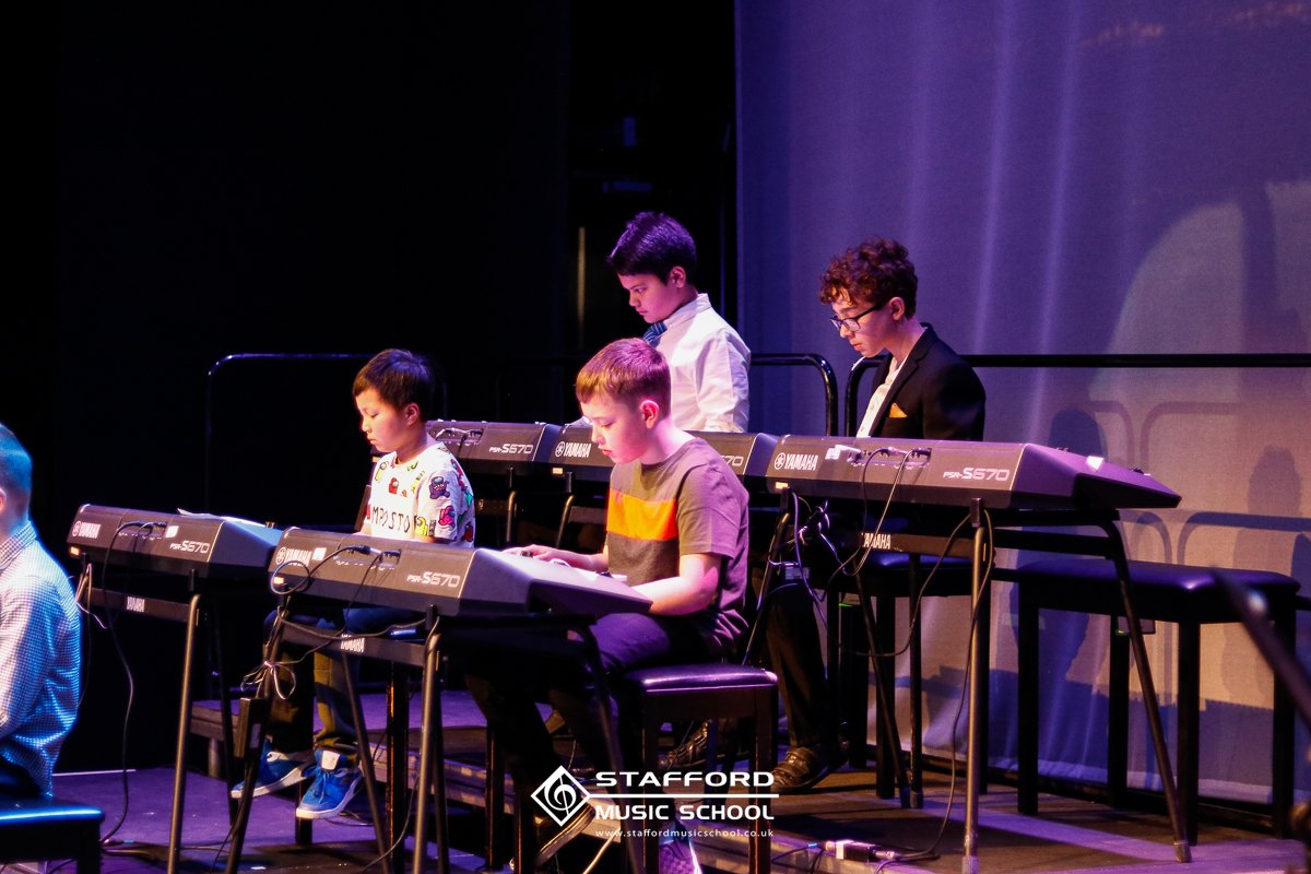 Group of children playing Yamaha keyboards on stage during a music performance at Stafford Music School.