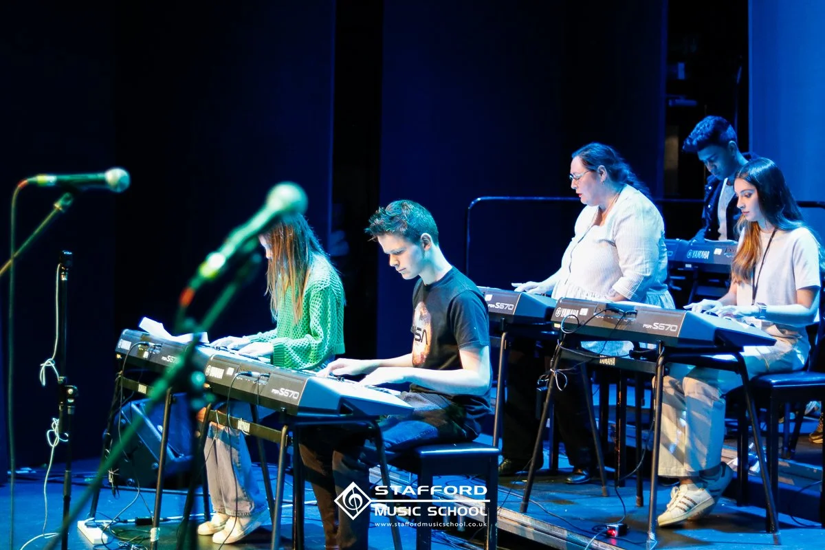 Five young musicians, three females and two males, are playing electronic keyboards on stage during a performance. They are focused on their instruments under stage lighting with a dark background. The logo of Stafford Music School is visible at the 