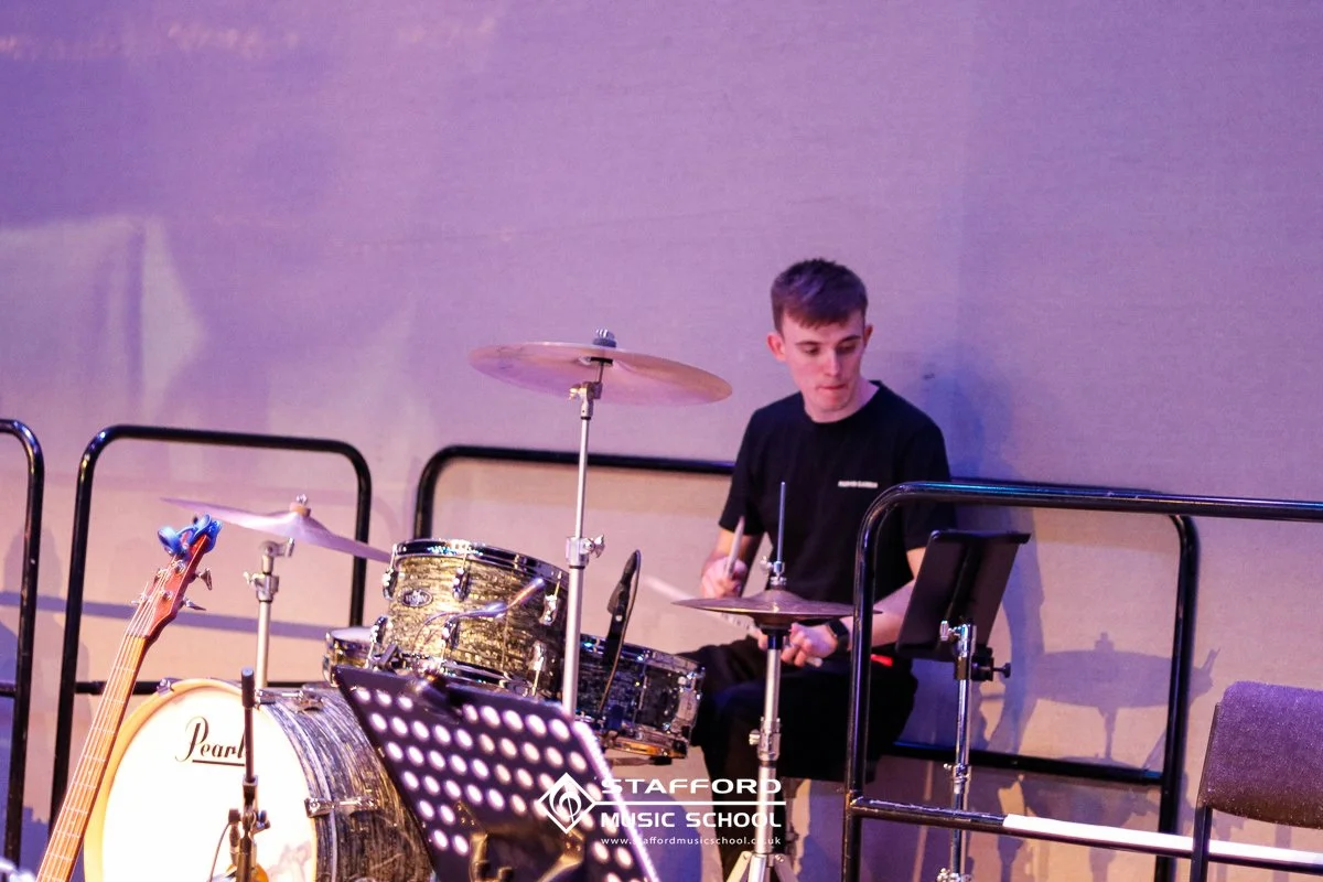A young man playing drums on stage at Stafford Music School, with a guitar resting against the drum set.