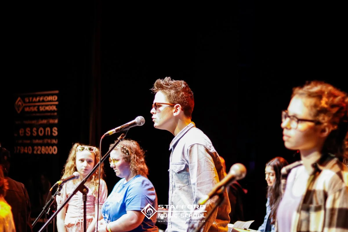 A group of young people standing on a stage with microphones during a performance or rehearsal at Stafford Music School, with a black background.