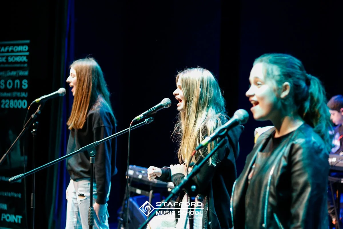Three young women singing on stage with microphones, in a music school concert setting, with a keyboard in the background, and a sign reading 'Stafford Music School' in the foreground.
