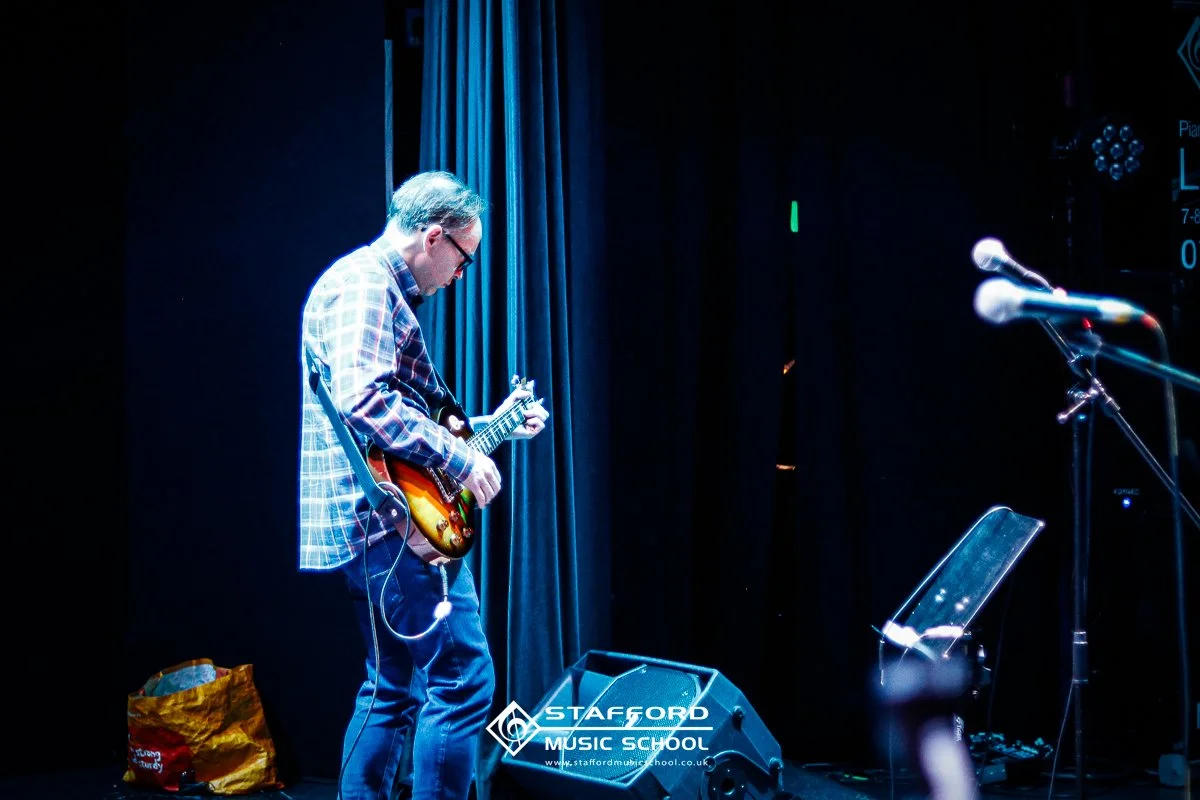 A man playing an electric guitar on stage with dark curtains behind him, microphone stands to the right, and a music stand in front of him. The stage floor has a yellow bag and a speaker. The logo for Stafford Music School is visible at the bottom.