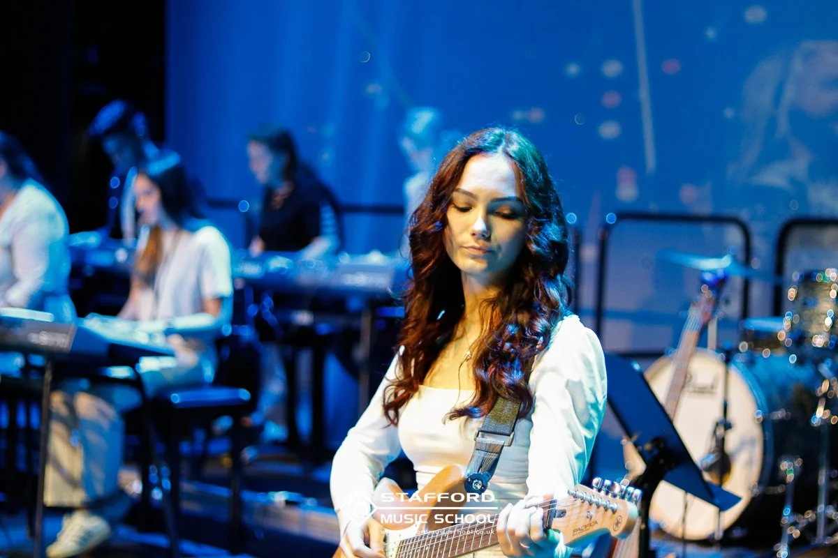 Young woman with long wavy hair playing electric guitar on stage with a band in the background, stage lights, and musical instruments.