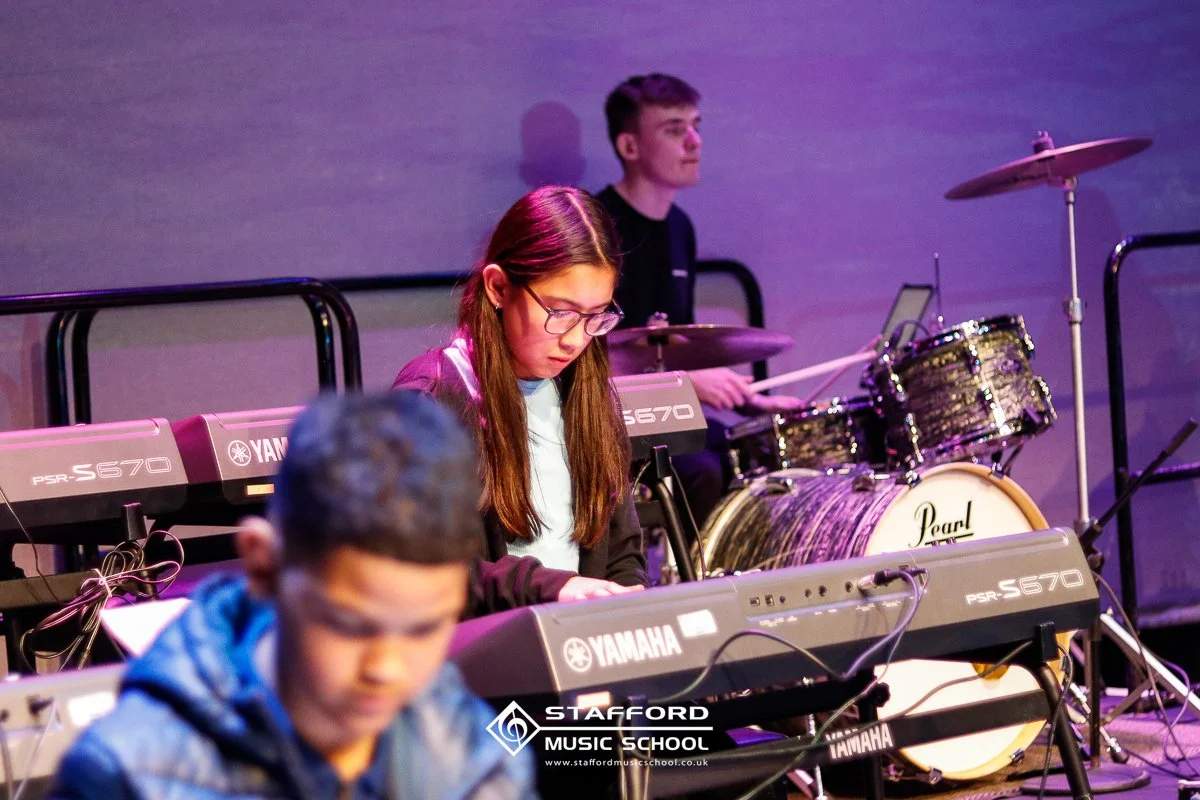 Young girl playing keyboard with a boy playing drums in the background during a music performance at Stafford Music School.