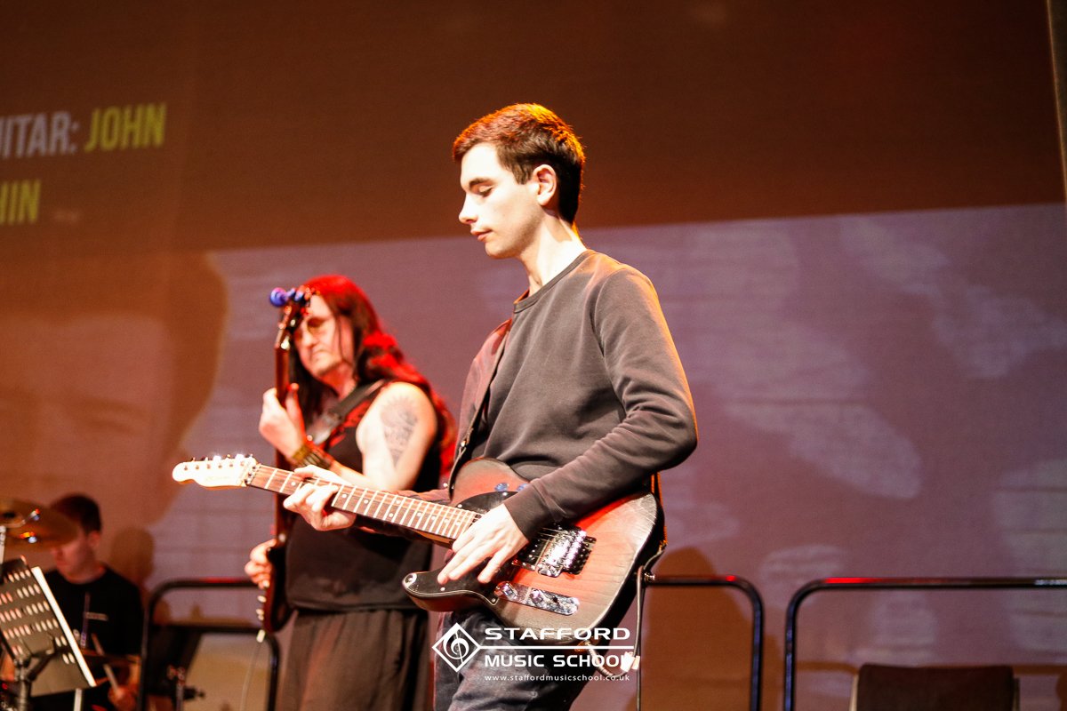 Two young musicians practicing guitar on stage at Stafford Music School. One male and one female, with music stands visible in the background.