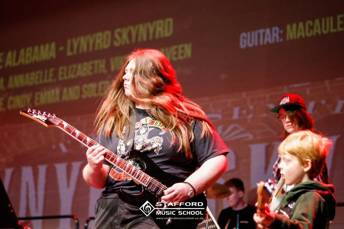 Young woman with long hair playing an electric guitar on stage at Stafford Music School, with other musicians and a projected screen in the background.