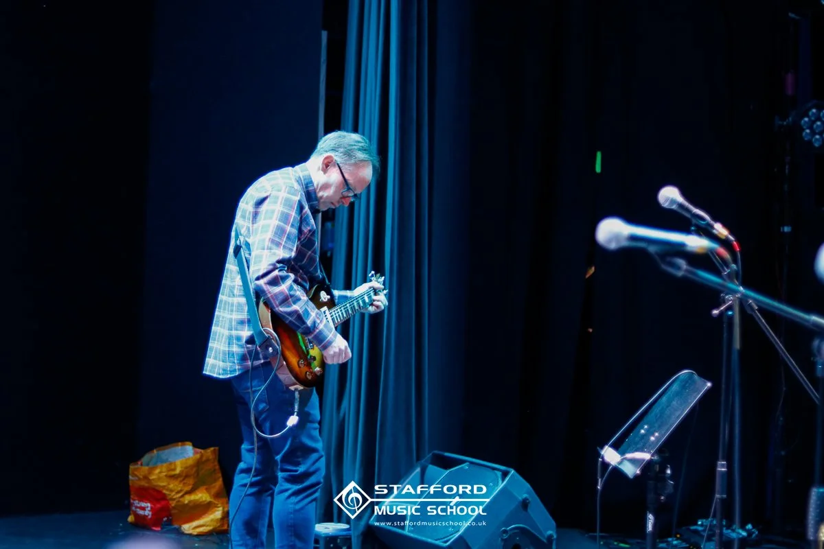 A man in a plaid shirt and glasses is playing an electric guitar on stage with blue curtains behind him and microphones nearby.