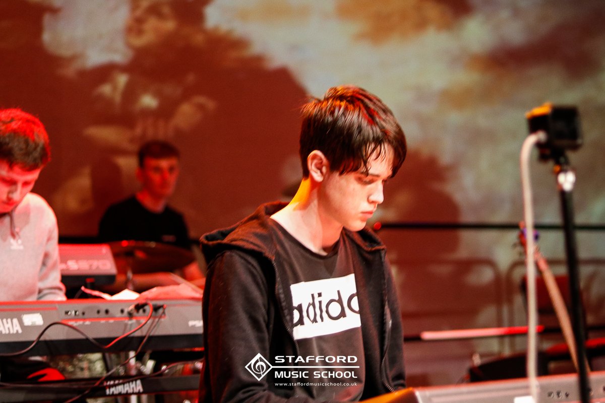 Young man with dark hair, wearing a black Adidas shirt and a hoodie, standing in a music rehearsal space with electronic keyboards and a drummer in the background.