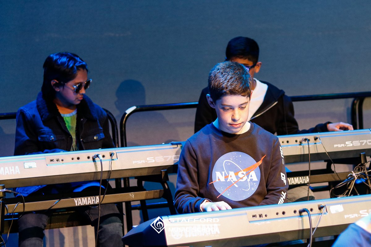 Three young people playing electronic keyboards on a stage; the boy in the foreground is wearing a NASA sweatshirt, and the two in the background are wearing glasses and dark clothing.