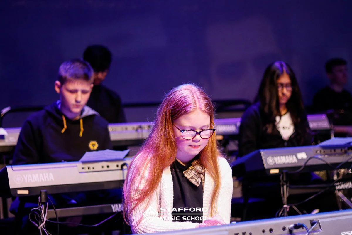 A group of young students playing Yamaha keyboards during a music performance, with a girl with red hair and glasses in the front, and others in the background.