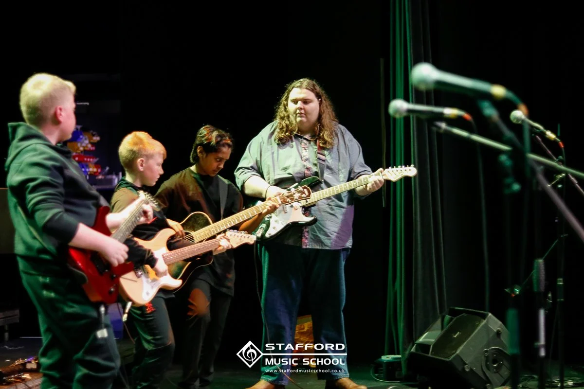 Four young people on stage playing guitars, one with a microphone in front of them, at Stafford Music School, with microphones and stage equipment visible.