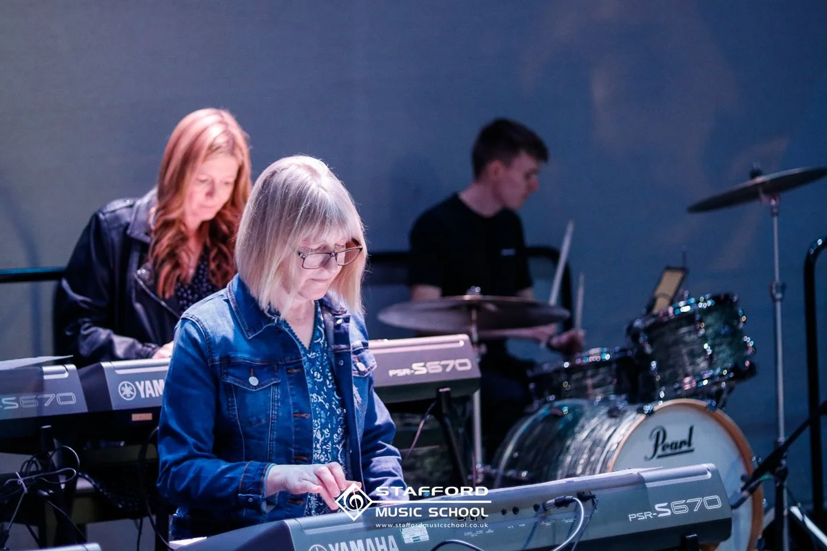 Three musicians playing instruments on stage: a woman with glasses and blonde hair playing a Yamaha keyboard, a woman with red hair playing a Yamaha keyboard, and a young man in the background playing a drum set.